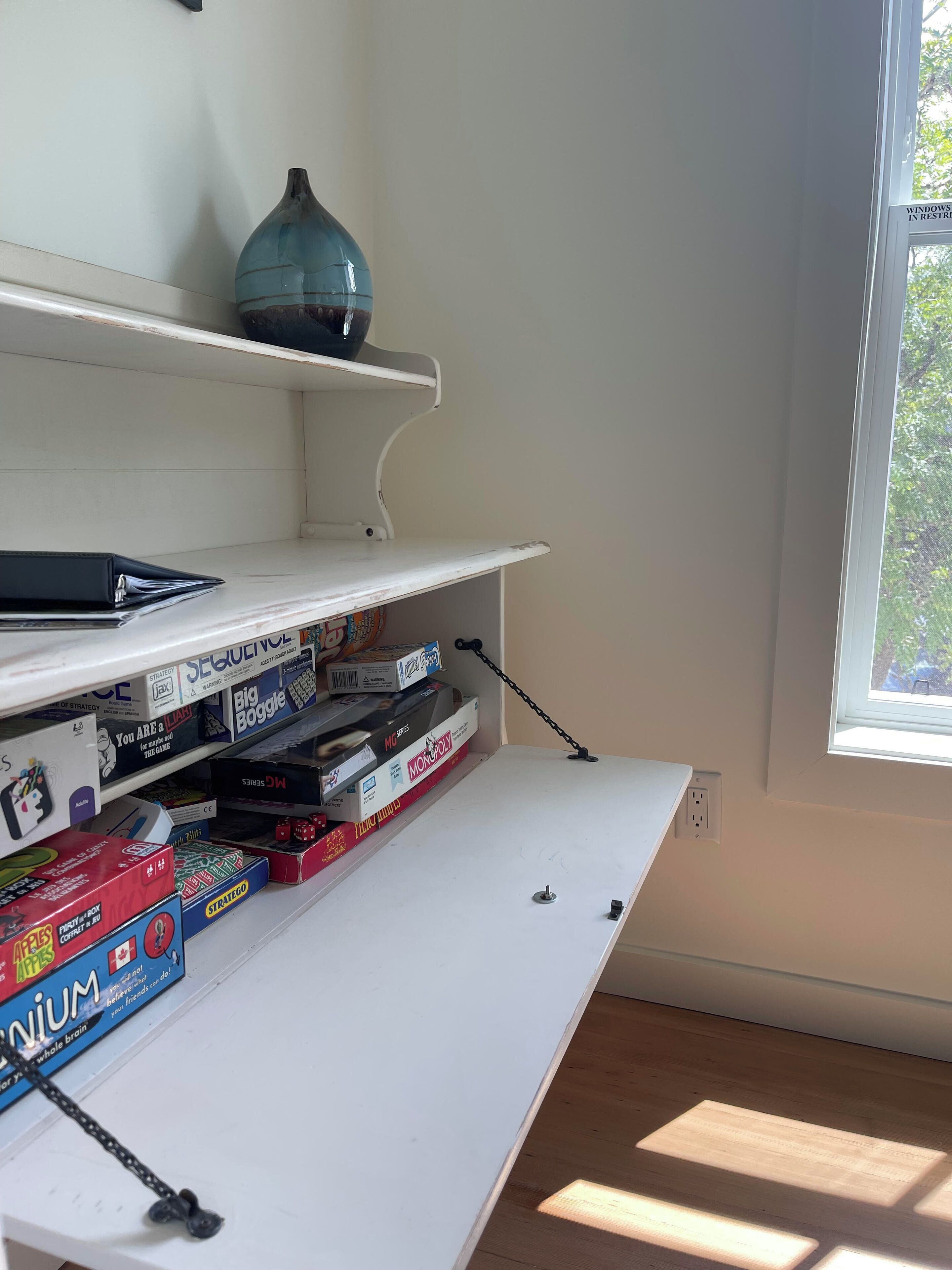 A room with a white desk, shelves with board games, a vase, and a window with a view of trees.