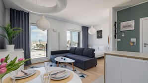 Un salón moderno con un sofá gris, una mesa de comedor blanca con platos y vasos, una mesita auxiliar blanca, y vista a la ciudad a través de amplios ventanales.