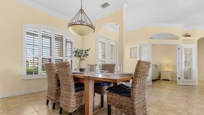 A dining area with a wooden table and wicker chairs, large windows with shutters, and a hanging light fixture. - Gulf Access, Kayaks, Heated Pool - Villa Happy Hour - Roelens Vacations (Cape Coral)