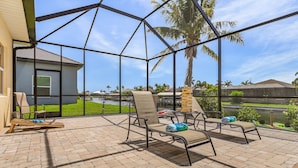 A covered patio with lounge chairs and a table, overlooking a residential area with palm trees and a body of water. - Gulf Access, Kayaks, Heated Pool - Villa Happy Hour - Roelens Vacations (Cape Coral)