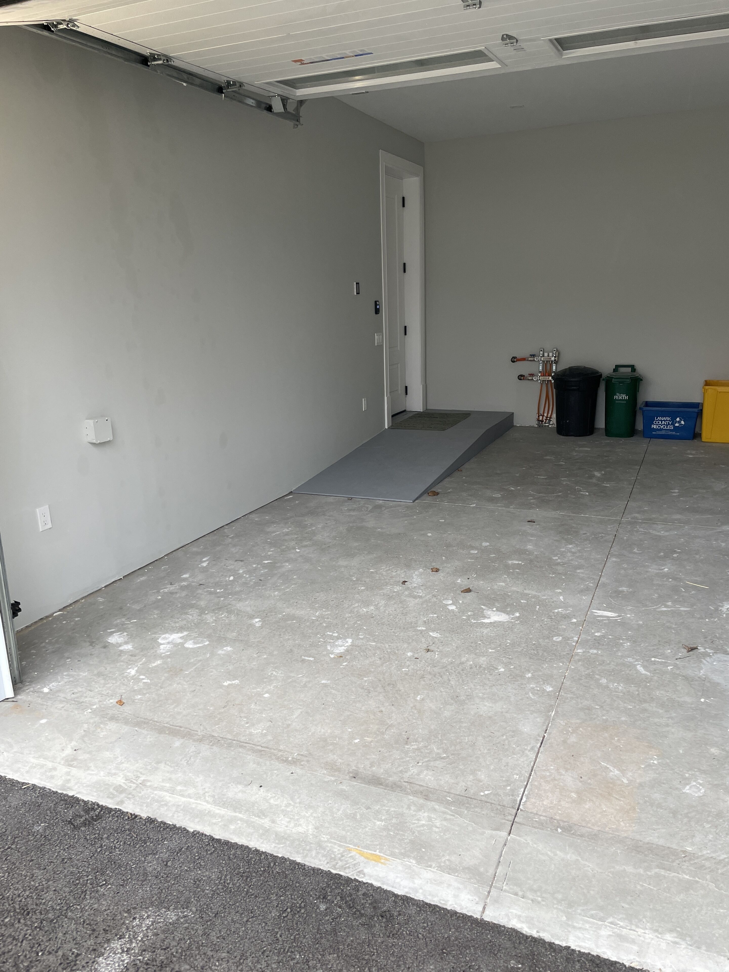 En garage med betongulv, en hvid dør og en lille rampe, der fører op til døren. Der er to skraldespande og en genbrugsbeholder op ad væggen.