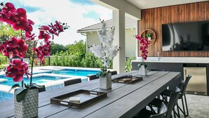 Un espace extérieur aménagé pour les repas, avec une piscine, un mur en bois servant de support pour un téléviseur et des plantes orchidées.
