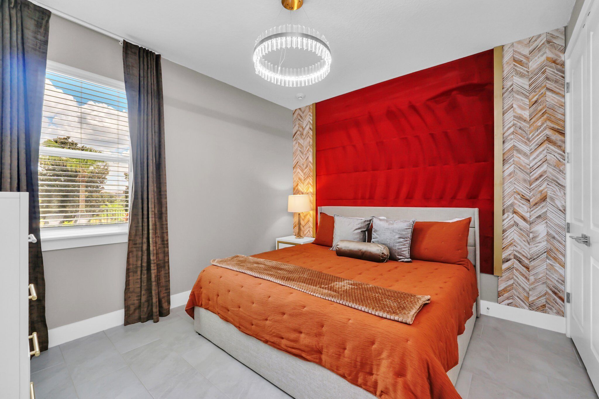 Une chambre avec un grand lit, un lustre, une fenêtre avec des rideaux et un mur d’accent rouge.