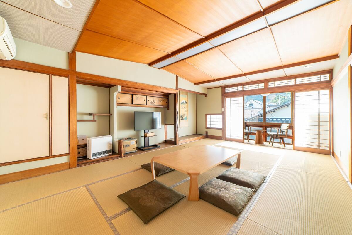 Una habitación japonesa tradicional con suelo de tatami, una mesita baja, cojines, puertas correderas y vistas al exterior.