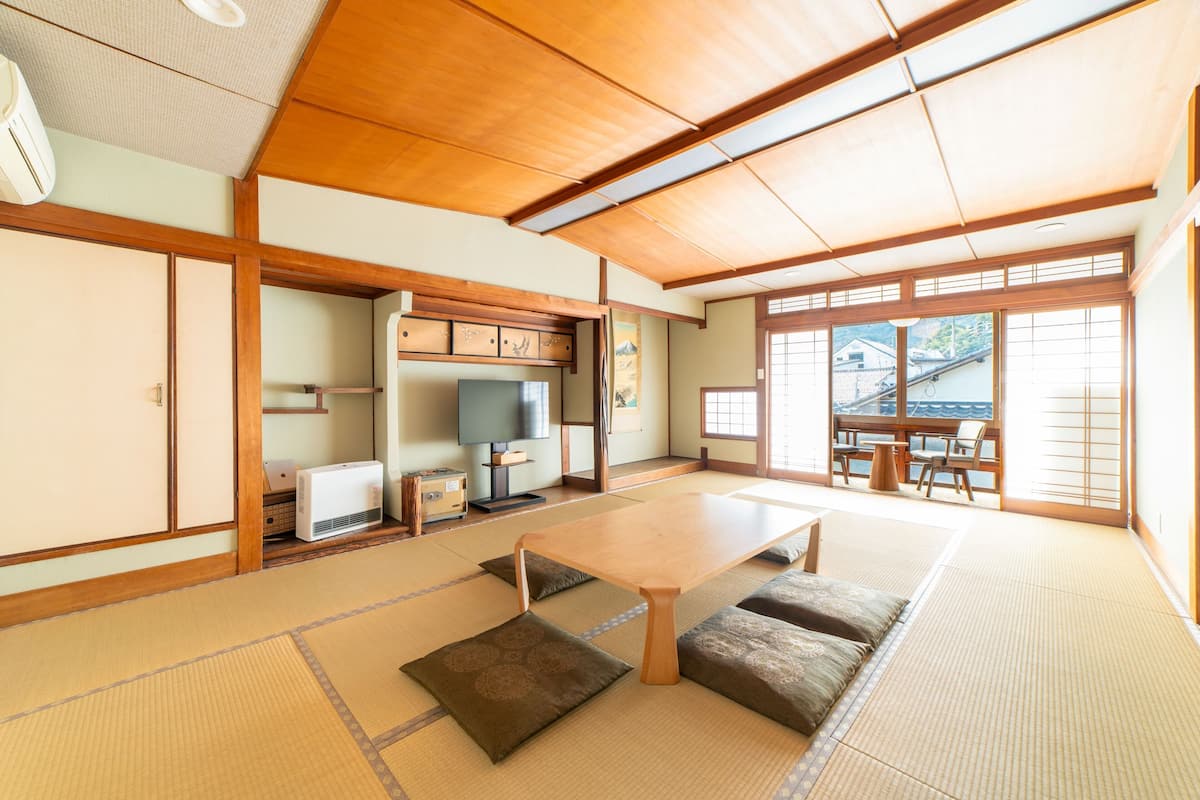 A traditional Japanese room with tatami flooring, a low table, cushions, sliding doors, and a view of the outside.