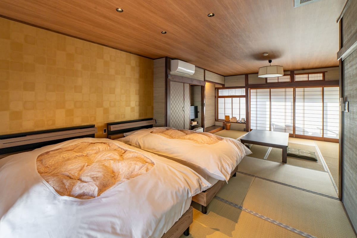 Una habitación de estilo japonés tradicional con suelo de tatami, dos camas con ropa blanca, una mesita y vistas a través de biombos shoji.