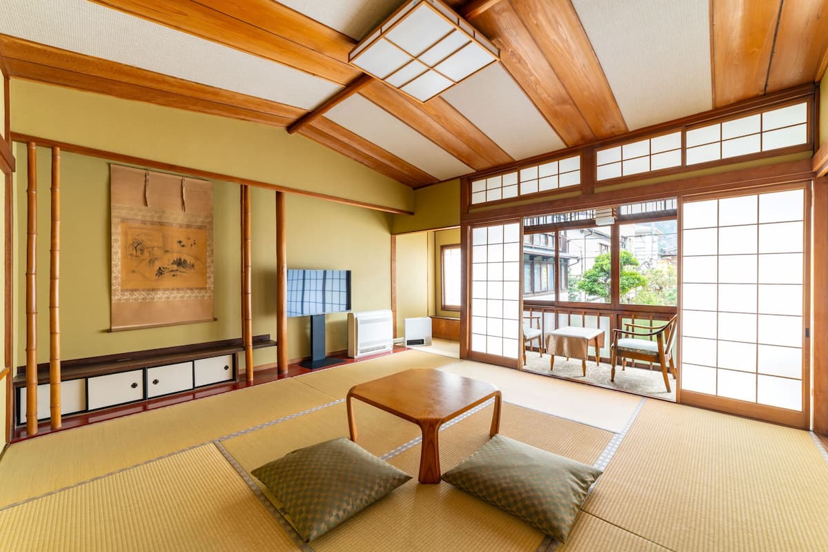 Una habitación japonesa tradicional con suelo de tatami, una mesa de madera baja, cojines, puertas correderas y vistas al exterior.