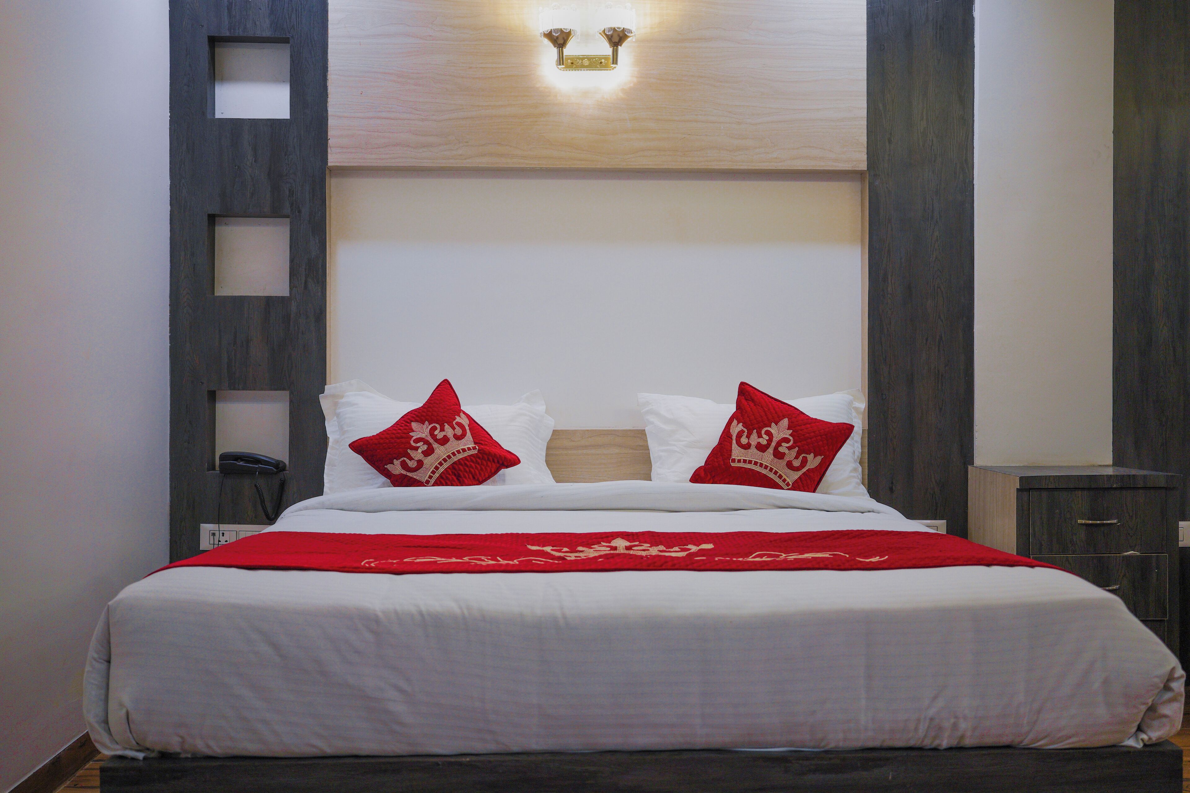 A neatly made bed with white linens and red pillows, a bedside telephone, and a wooden headboard with a light fixture above.