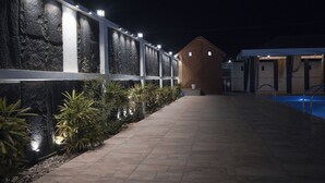 Vue nocturne d’un couloir d’hôtel avec une piscine, un mur de pierre illuminé et un bâtiment en briques en arrière-plan.
