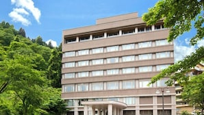 Um hotel com vários andares, uma entrada central, cercado por vegetação e um acesso pavimentado.