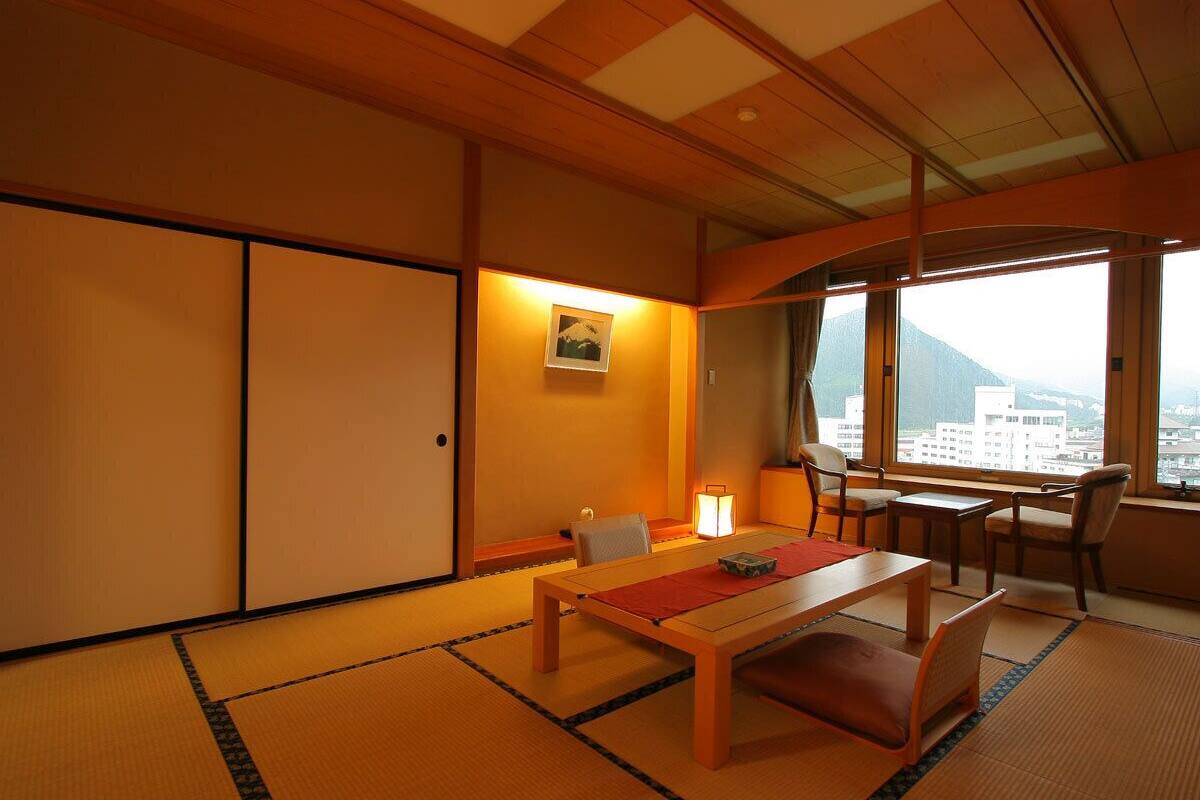 Ein traditioneller japanischer Raum mit Tatami-Bodenbelag, einem niedrigen Tisch, Stühlen, einer Schiebetür und Blick auf eine Stadtlandschaft.