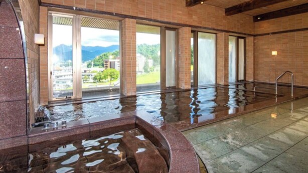 Uma ampla piscina coberta com paredes de vidro transparente, piso de pedra e vista para montanhas e edifícios lá fora.
