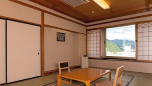 Um quarto japonês tradicional com tatames, uma mesa de madeira baixa, duas cadeiras, portas deslizantes, um aquecedor e vista para montanhas por grandes janelas.