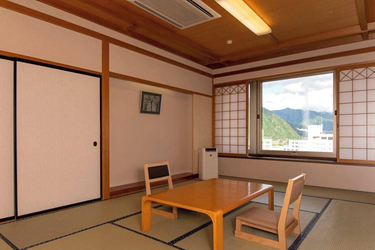 Ein traditioneller japanischer Raum mit Tatami-Böden, einem niedrigen Holztisch, zwei Stühlen, Schiebetüren, einer Heizung und einer Aussicht auf Berge durch große Fenster.
