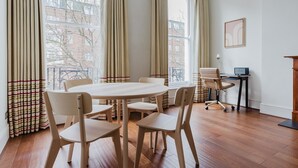 Ein Zimmer mit einem runden Holztisch, vier Stühlen, einem Schreibtisch mit Computer und großen Fenstern mit Vorhängen.