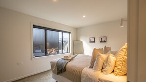 Un dormitorio con un ventanal grande, una cama con múltiples almohadas, una mesita de noche y dos cuadros en la pared.