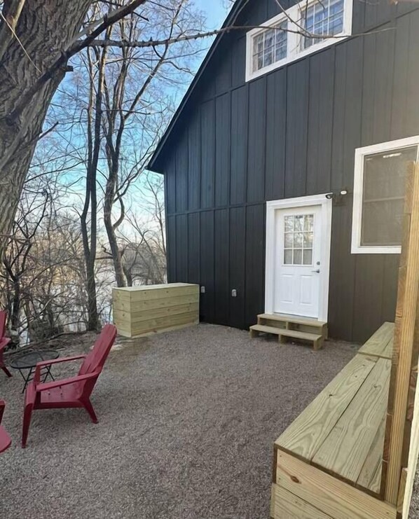 A dark wooden building with a white door and windows, a gravel patio, and red chairs.