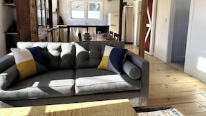 A cozy living room with a grey sofa, wooden beams, a dining area, and a kitchen in the background. - Private Riverfront, Converted Barn *EV Charger* (Princeton)