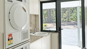 A compact laundry room with a front-loading washer and dryer, a sink, and a window with a view of a wooded area. - Springdale Gums: Denmark Forest Retreat (Hay)