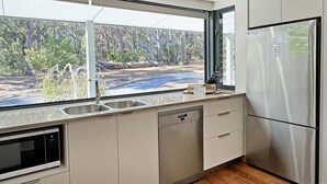 A modern kitchen with a built-in dishwasher, a stainless steel refrigerator, and a large window offering a view of a forested area. - Springdale Gums: Denmark Forest Retreat (Hay)