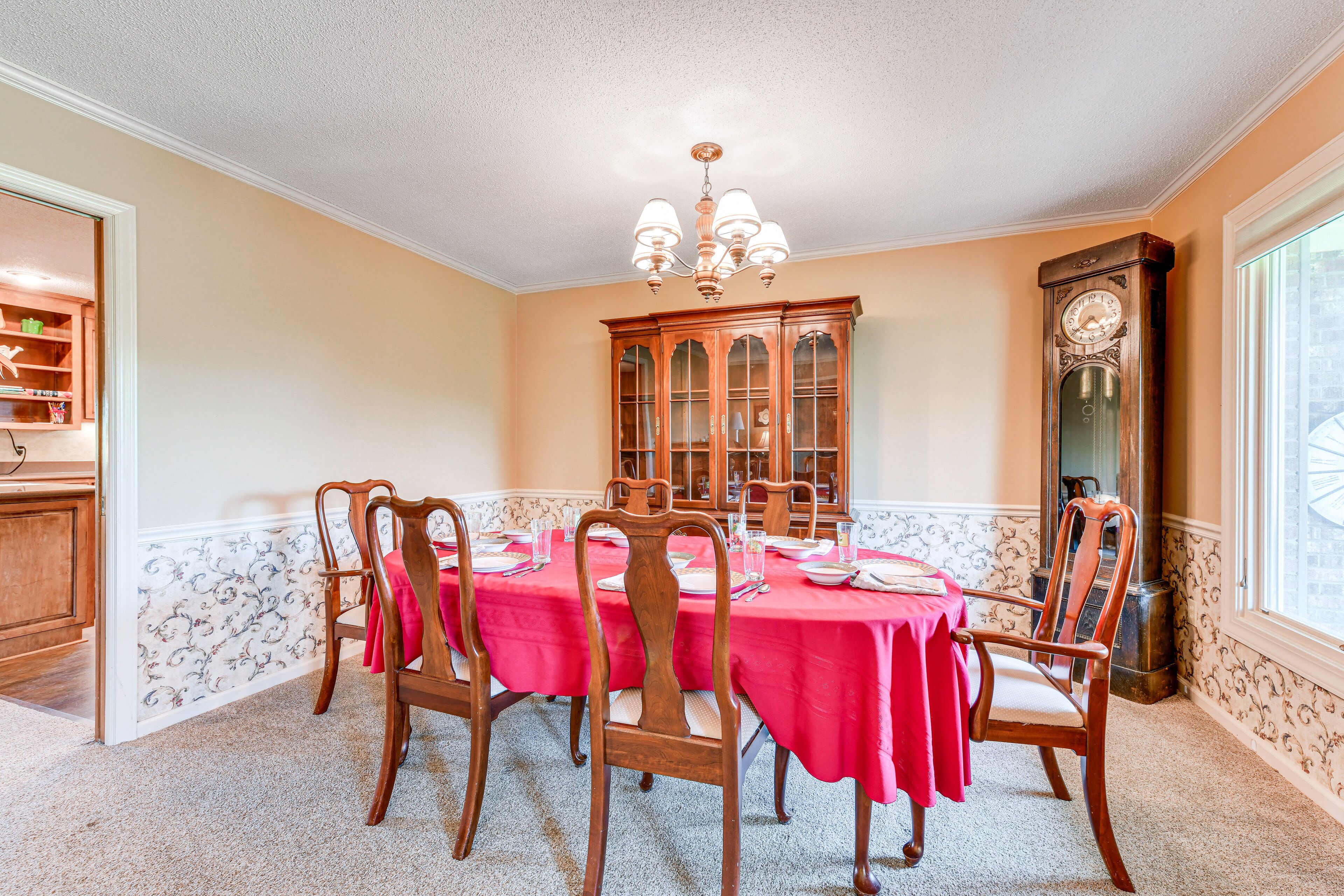 Uma sala de jantar com uma mesa redonda posta para seis pessoas, rodeada por cadeiras de madeira, um candelabro e um armário de madeira grande com portas de vidro.