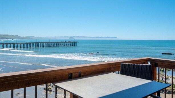 A beach view with a wooden deck, a table, and a view of the ocean and a pier in the distance. - Oceanfront condo with PRIVATE deck. Pet friendly. Just a short walk to town. (Cayucos)