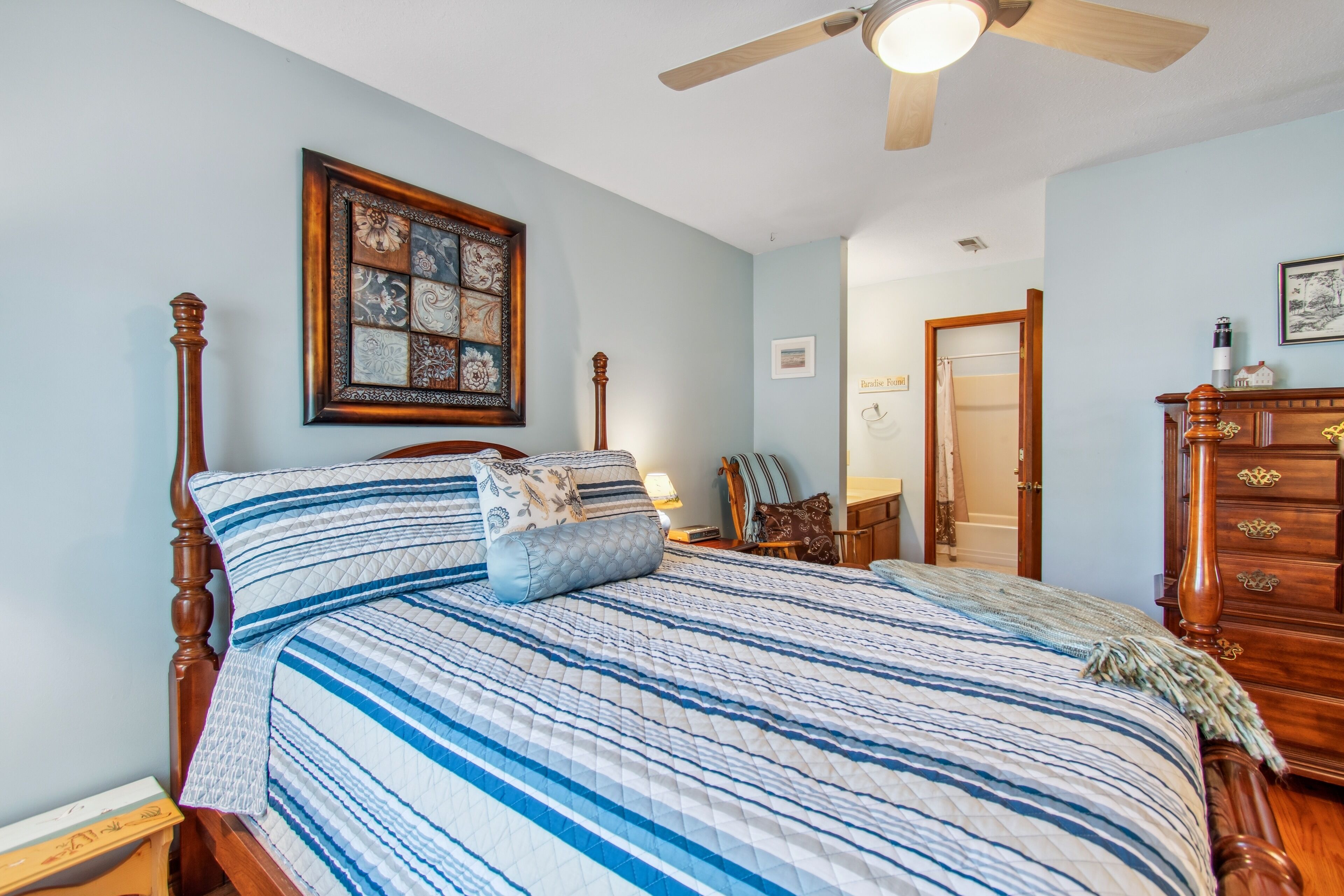 A bedroom with a large bed, a ceiling fan, a wooden dresser, and a framed artwork on the wall.