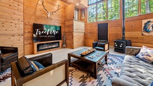 A log cabin interior with a fireplace, a large TV, leather furniture, and a large window with a view of trees. - The Roundhouse Lodge | Pet-Friendly Game Retreat (Broken Bow)