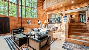 A spacious living area with a large window, a fireplace, and a staircase leading to an upper level. - The Roundhouse Lodge | Pet-Friendly Game Retreat (Broken Bow)