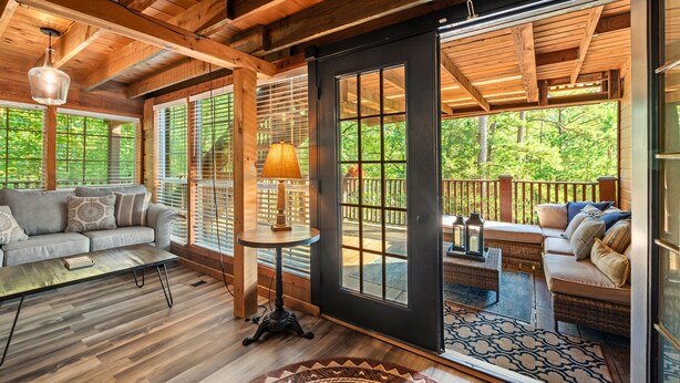 A cozy cabin interior with a wooden floor, a sofa, a table, and a large sliding glass door leading to a deck. - The Roundhouse Lodge | Pet-Friendly Game Retreat (Broken Bow)