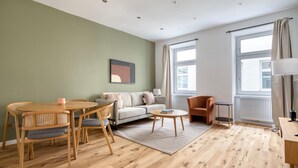A modern living room with a wooden floor, a grey sofa, a round dining table with chairs, a small side table, and a floor lamp.
