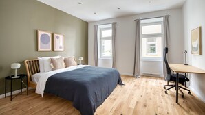 A modern bedroom with a large bed, a desk, and two windows with curtains.