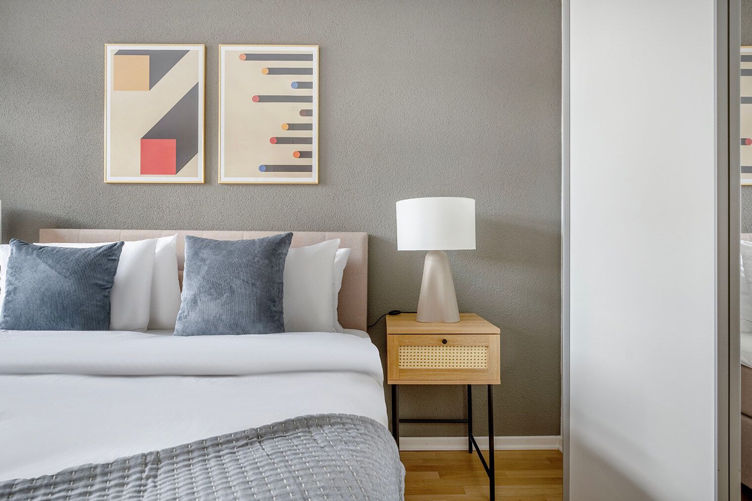 Een netjes opgemaakte slaapkamer met een bed, kussens, een nachtkastje met een lamp en twee abstracte kunstwerken aan de muur.