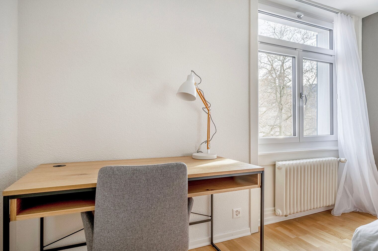 A room with a wooden desk, a grey chair, a white radiator, and a window with white curtains.