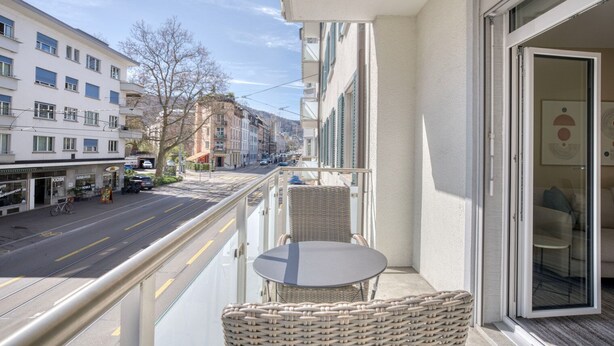 Un balcon avec une chaise et une table en osier, donnant sur une rue bordée d’immeubles et traversée par une ligne de tramway.