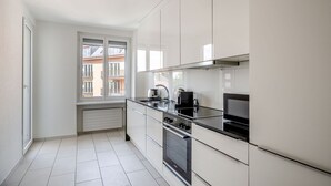 Une cuisine moderne avec des meubles blancs, un plan de travail noir, une cuisinière et une fenêtre donnant sur des immeubles.