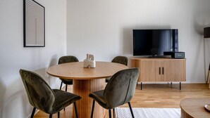 Un salon moderne avec une table de salle à manger ronde en bois, quatre chaises rembourrées, un téléviseur posé sur un meuble en bois et une œuvre d'art abstraite encadrée accrochée au mur.