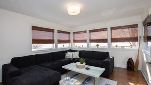 Ein modernes Wohnzimmer mit einem schwarzen Ecksofa, einem weißen Couchtisch mit Pflanze und großen Fenstern mit Jalousien.