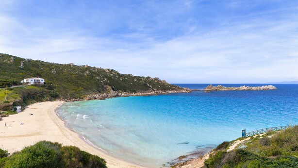 A coastal landscape with a sandy beach, clear blue waters, and a rocky outcrop.