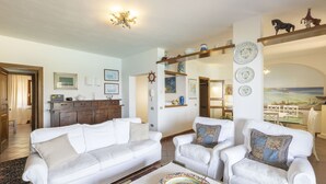 A spacious living room with a white sofa, armchairs, and a coffee table. There is a kitchen area in the background with wooden cabinets and a dining table.