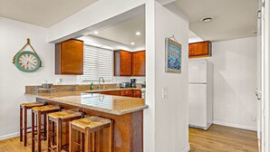 A kitchen with a central island, wooden bar stools, wooden cabinets, a white refrigerator, and a clock on the wall.