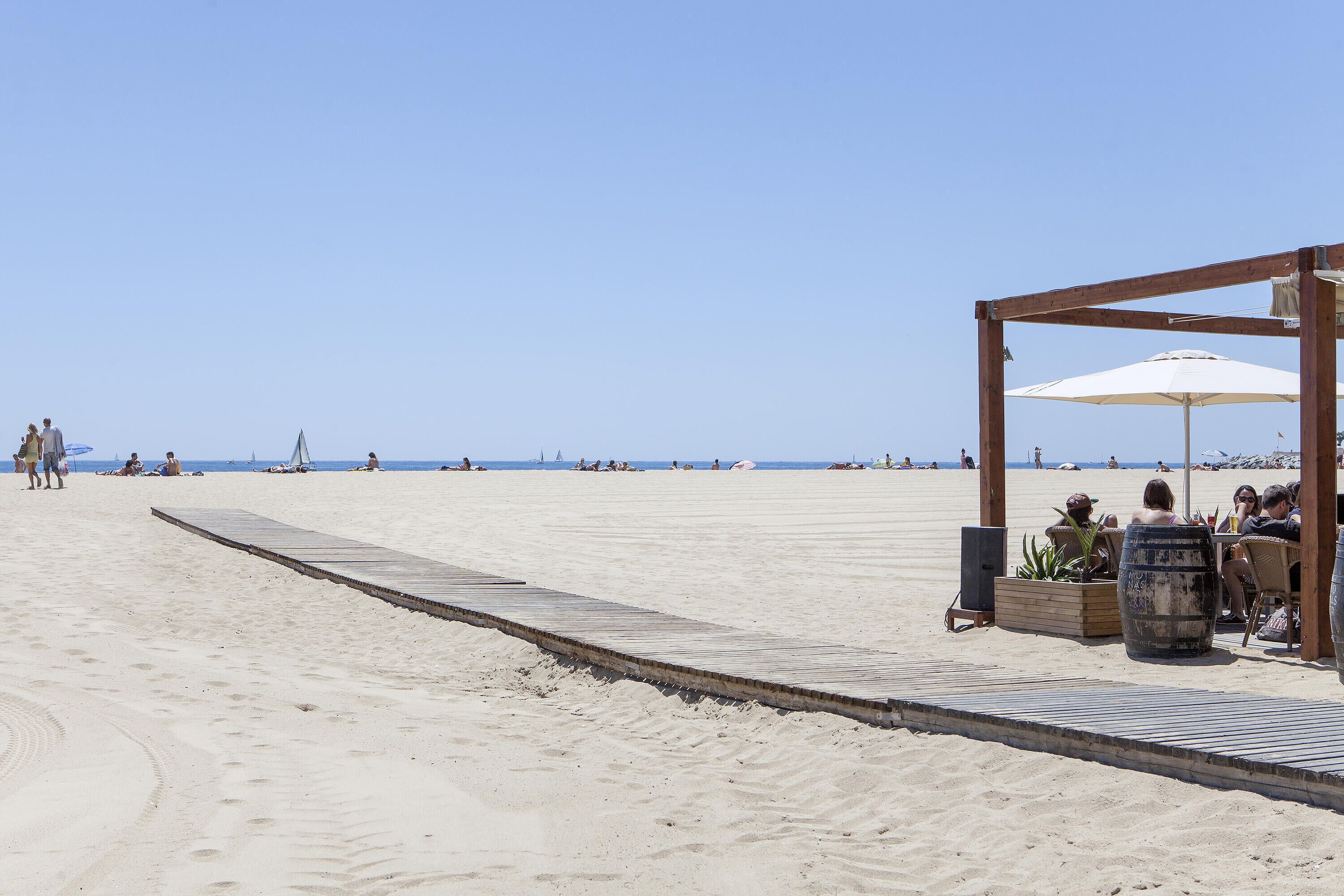 Una estructura de madera frente a la playa con una sombrilla blanca, zona de estar y un macetero de madera.