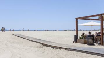Una estructura de madera frente a la playa con una sombrilla blanca, zona de estar y un macetero de madera.