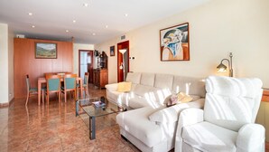Sala de estar com sofá, poltrona e mesa de centro de vidro. Há também um espaço de jantar com mesa e cadeiras.
