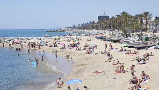 Uma praia movimentada com pessoas se bronzeando e nadando, e barcos alinhados na orla.
