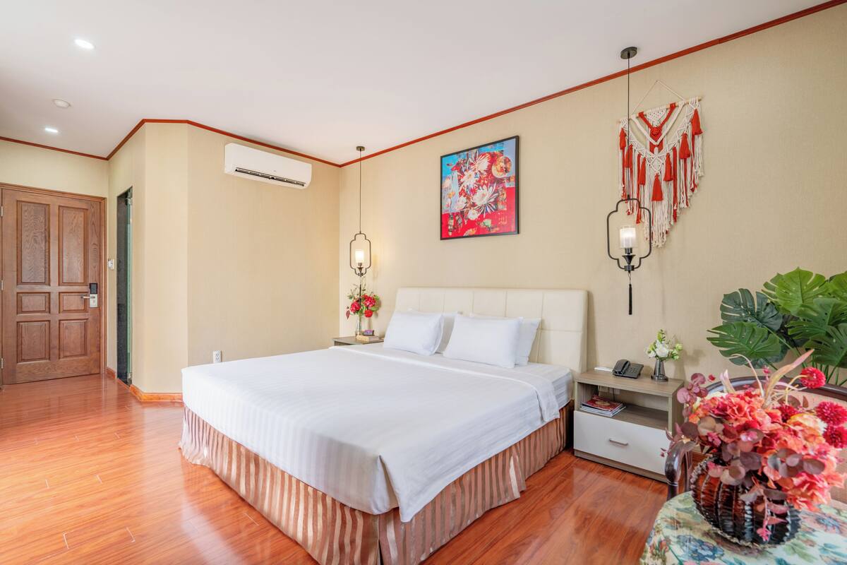 A bedroom with a large bed, a bedside table with a lamp, a wall-mounted air conditioner, and a decorative macrame piece.