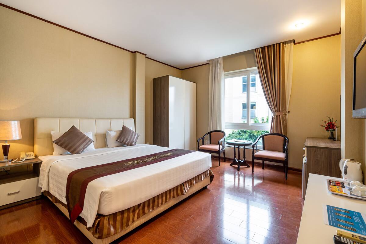 A hotel room with a large bed, bedside table with a lamp, a chair, a small table with a kettle and cups, and a window with curtains.