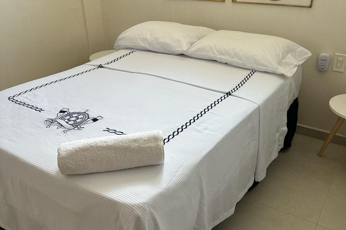 Een net opgemaakt bed met een witte spreidek en een decoratief kussen. Daarboven hangen twee decoraties aan de muur.