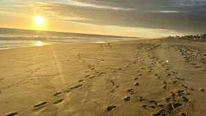 Una playa con huellas que conducen al agua, un atardecer en el fondo y un cielo nublado.