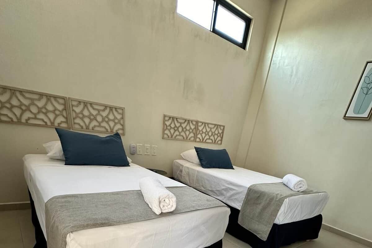 Een hotelkamer met twee bedden, elk met een witte sprei, een grijze deken en een opgerolde witte handdoek. De kamer heeft een betegelde vloer en een raam met uitzicht op het exterieur.